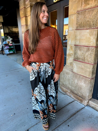 A woman stands on a sidewalk wearing a rust-orange long-sleeve knit top with a white eyelet pattern and black wide-leg pants featuring a large white and gray tropical leaf print with brown accents.