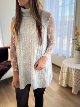 Person wearing a long, light-colored sweater with lace details in a room with large windows.