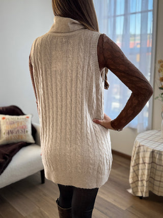 Person wearing a beige knitted vest in a room with furniture and decor.