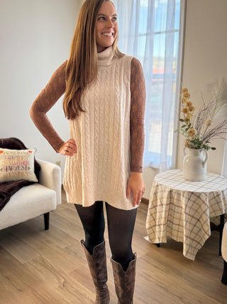 Woman wearing a beige turtleneck dress and gray knee-high boots in a living room.
