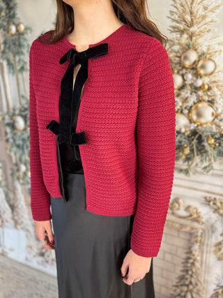 Person wearing a red knitted cardigan with a black bow tie against a decorative background.