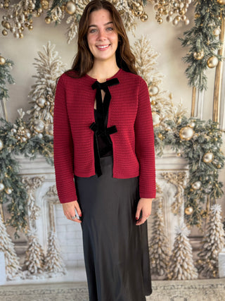 Woman wearing a red jacket with a black bow tie in front of a decorated Christmas background.