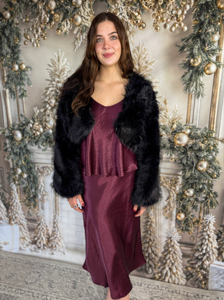 Woman in a burgundy dress with a black fur coat standing in front of a decorated Christmas background.