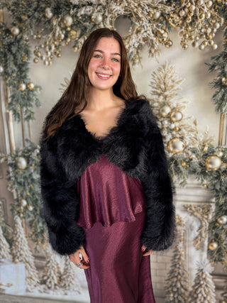 Woman wearing a black fur shrug over a burgundy dress in front of a decorated Christmas background.