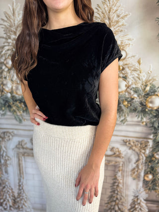 Woman wearing a black velvet top and white skirt against an ornate wall.