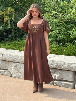 A woman wearing a brown midi dress with short puff sleeves and a decorative embroidered bodice, standing outdoors in front of a stone wall and green foliage.