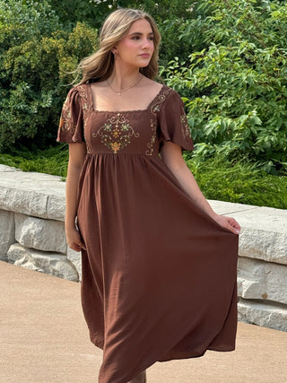 A woman wearing a brown midi dress with short puff sleeves and an embroidered bodice, standing outdoors in front of a stone wall and green foliage.