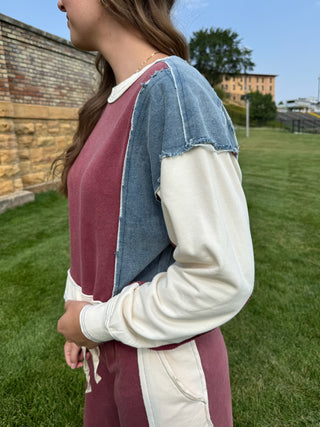 A woman wearing a color-blocked sweatshirt with a burgundy body, a blue denim shoulder panel, and off-white sleeves, standing outdoors on a grassy lawn.