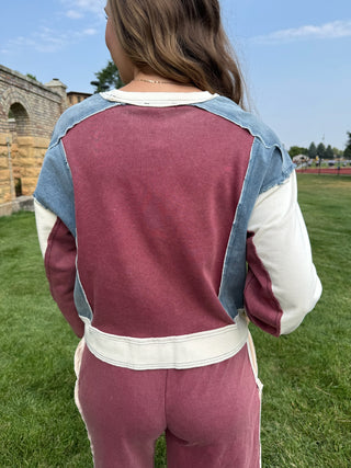 A person viewed from behind wearing a color-blocked sweatshirt and matching sweatpants set. The set features a dusty rose main body with light blue denim shoulder panels and off-white sleeve and waist accents, set against a grassy field with stone ruins in the background.
