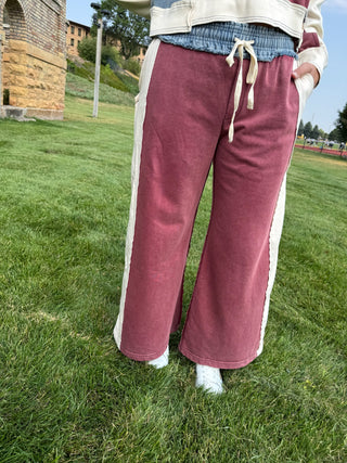 A person standing outdoors on grass wearing wide-leg sweatpants in a dusty rose color with cream side panels and drawstrings, paired with a blue denim waistband and a cream jacket.