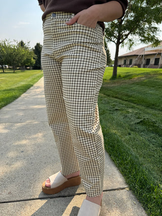 A person outdoors wearing beige and brown gingham check pants, standing on a concrete path with grass and trees in the background.
