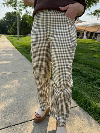 A person outdoors wearing beige and brown gingham check pants, standing on a concrete path with grass and trees in the background.