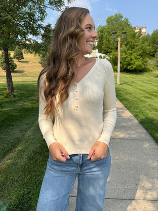 A woman wearing a cream ribbed long-sleeve henley sweater and blue jeans, standing on a concrete path in a grassy park area.