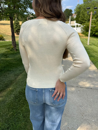 A woman wearing a cream ribbed knit long-sleeve top and light blue jeans, photographed from behind outdoors on a paved path with grass and trees in the background.