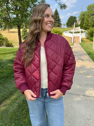 A woman wearing a burgundy quilted bomber jacket, a cream ribbed knit top, and light blue jeans, standing outdoors on a paved path with trees and grass in the background.