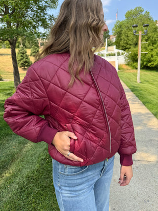 A woman wearing a quilted bomber jacket in burgundy, standing outdoors on a paved path with grass and trees in the background.
