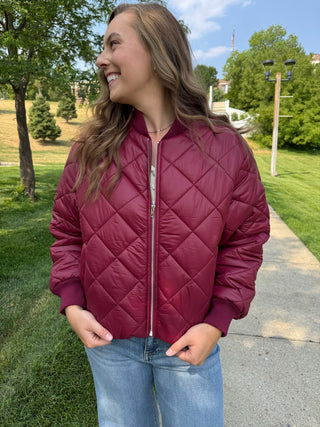 A woman wearing a burgundy quilted bomber jacket and blue jeans, standing outdoors on a paved path in a park with green grass and trees.