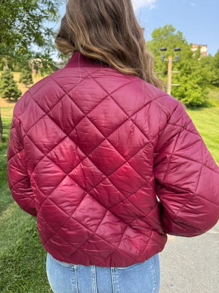 A person viewed from behind wearing a quilted bomber-style jacket in a deep berry red or burgundy color, standing outdoors on a paved path with grass, trees, and blue sky in the background.