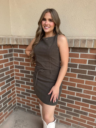 A woman wearing a sleeveless dark brown corduroy mini dress with a matching belt, paired with white cowboy boots, posing in front of a brick wall.