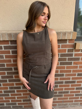 A woman wearing a sleeveless dark brown mini dress with a matching tie belt, standing against a brick wall.