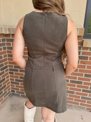 A woman viewed from behind wearing a sleeveless, knee-length brown dress with a structured back and waist seam, standing outdoors against a brick and stucco wall.