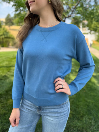 A woman wearing a light blue crewneck knit sweater and light wash jeans, standing outdoors on a grassy lawn with trees in the background.
