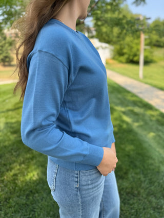 A woman wearing a light blue crewneck knit sweater and light wash jeans, standing outdoors on a grassy lawn with trees in the background.