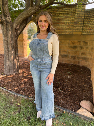 A woman wearing light-wash blue denim overall with a cream ribbed long-sleeve top, standing outdoors in front of a brick wall and tree.