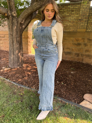 A woman wearing light-wash blue denim overall with a cream ribbed long-sleeve top and off-white pointed shoes, standing outdoors on grass next to a tree and brick wall.