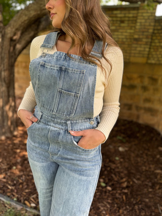 A woman wearing light-wash blue denim overall with a cream ribbed long-sleeve top, standing outdoors in front of a stone wall and tree.