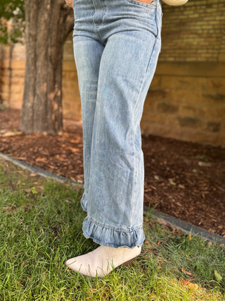 A person outdoors wearing light-wash blue flare jeans with a ruffled hem and off-white slip-on shoes, standing on grass near a tree and brick wall.