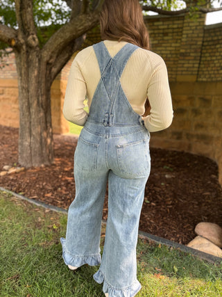 A woman viewed from behind wearing light blue denim overall with wide legs and ruffled hems, layered over a cream ribbed long-sleeve top, standing outdoors on grass near a tree and brick wall.