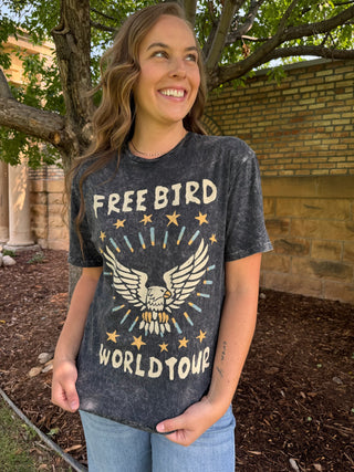 A woman wearing a dark gray acid-wash graphic t-shirt with a distressed eagle design and the text 'FREE BIRD WORLD TOUR' in cream and gold, standing outdoors in front of a brick wall and trees.