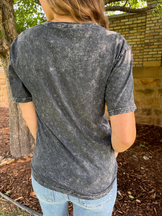 A person viewed from behind wearing a faded charcoal gray short-sleeve t-shirt and light blue jeans, standing outdoors in front of a brick wall and tree.
