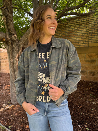 A woman wearing a faded gray-green camouflage utility jacket over a black graphic t-shirt and light blue jeans, standing outdoors in front of a brick wall and a tree.