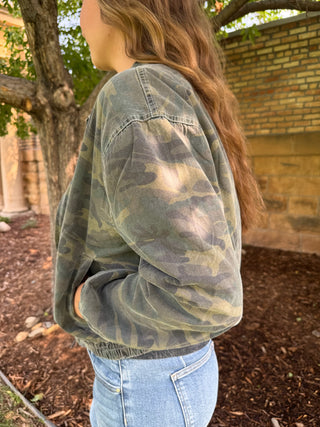 A woman wearing a washed camouflage pullover jacket in an outdoor setting with trees and a brick wall in the background.