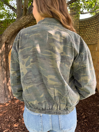 A woman viewed from behind wearing a faded camouflage jacket and light blue jeans, standing outdoors near a tree and brick wall.