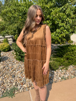 A woman wearing a sleeveless tiered brown dress with ruffle trim, standing outdoors on a concrete path with gravel and green bushes behind her.