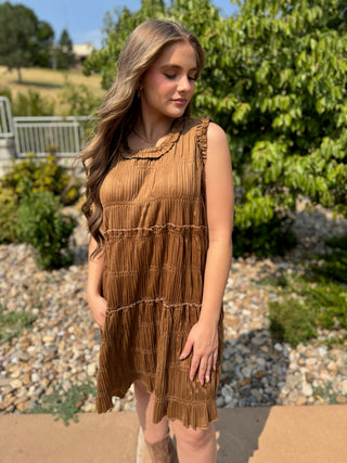 A woman wearing a sleeveless, tiered brown dress with a gathered, pleated texture, standing outdoors in front of green trees and a gravel path.