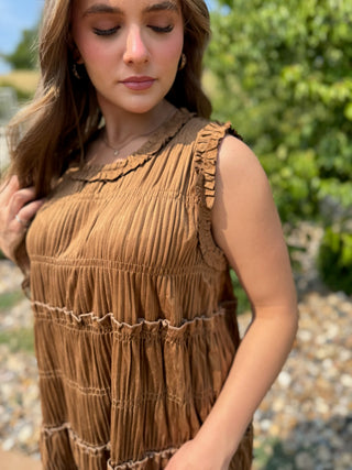 A woman wearing a sleeveless brown ruffled dress, photographed outdoors with green foliage and gravel in the background.