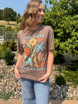 A woman wearing a brown graphic t-shirt with a colorful floral and bird design and the text 'IT IS WELL WITH MY SOUL', paired with light blue jeans, standing outdoors in a garden setting.