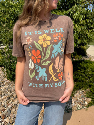 A woman wearing a brown acid-wash graphic t-shirt with colorful flowers and birds and the text 'IT IS WELL WITH MY SOUL', paired with blue jeans, standing outdoors in front of green foliage.