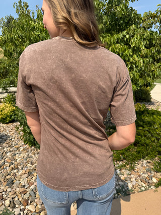 A woman viewed from behind wearing a brown acid-wash t-shirt and blue jeans, standing outdoors in a garden setting with green foliage and gravel.