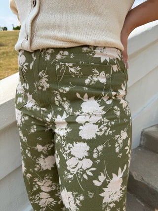 A close-up of a person wearing olive green pants with a white floral print, paired with a cream ribbed cardigan, standing outdoors next to a white wall.