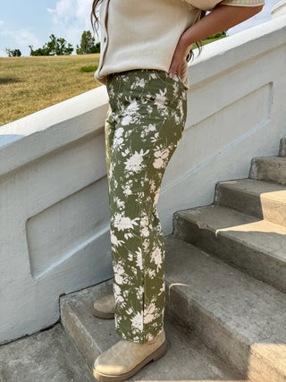 A person wearing olive green pants with a white floral print, standing on concrete outdoor stairs next to a white railing.