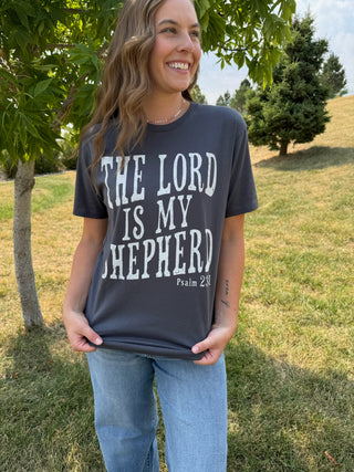 A woman wearing a dark charcoal gray graphic t-shirt with the text 'THE LORD IS MY SHEEPHERD Psalm 23' printed in white, standing outdoors on a grassy hill with trees in the background.