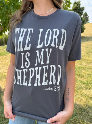 A woman wearing a dark charcoal gray graphic t-shirt with the text 'THE LORD IS MY SHEPHERD Psalm 23:1' printed in distressed white lettering, photographed outdoors on a grassy lawn.