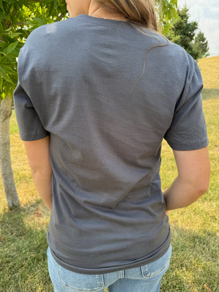 A person viewed from behind wearing a dark gray short-sleeve t-shirt and light blue jeans, standing outdoors in a grassy area with trees.