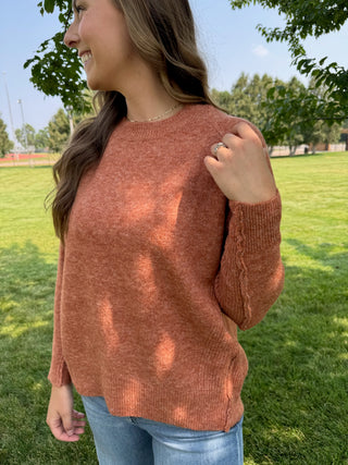 A woman wearing a heathered terracotta orange knit sweater and blue jeans, standing outdoors in a grassy park with trees in the background.