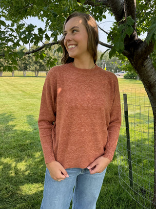 A woman wearing a heathered rust-orange long-sleeve knit sweater and light blue jeans, standing outdoors in a grassy park area under a tree.